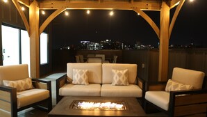 Un espacio exterior con asientos, un sofá y sillas, un hogar y vistas a la ciudad de noche.
