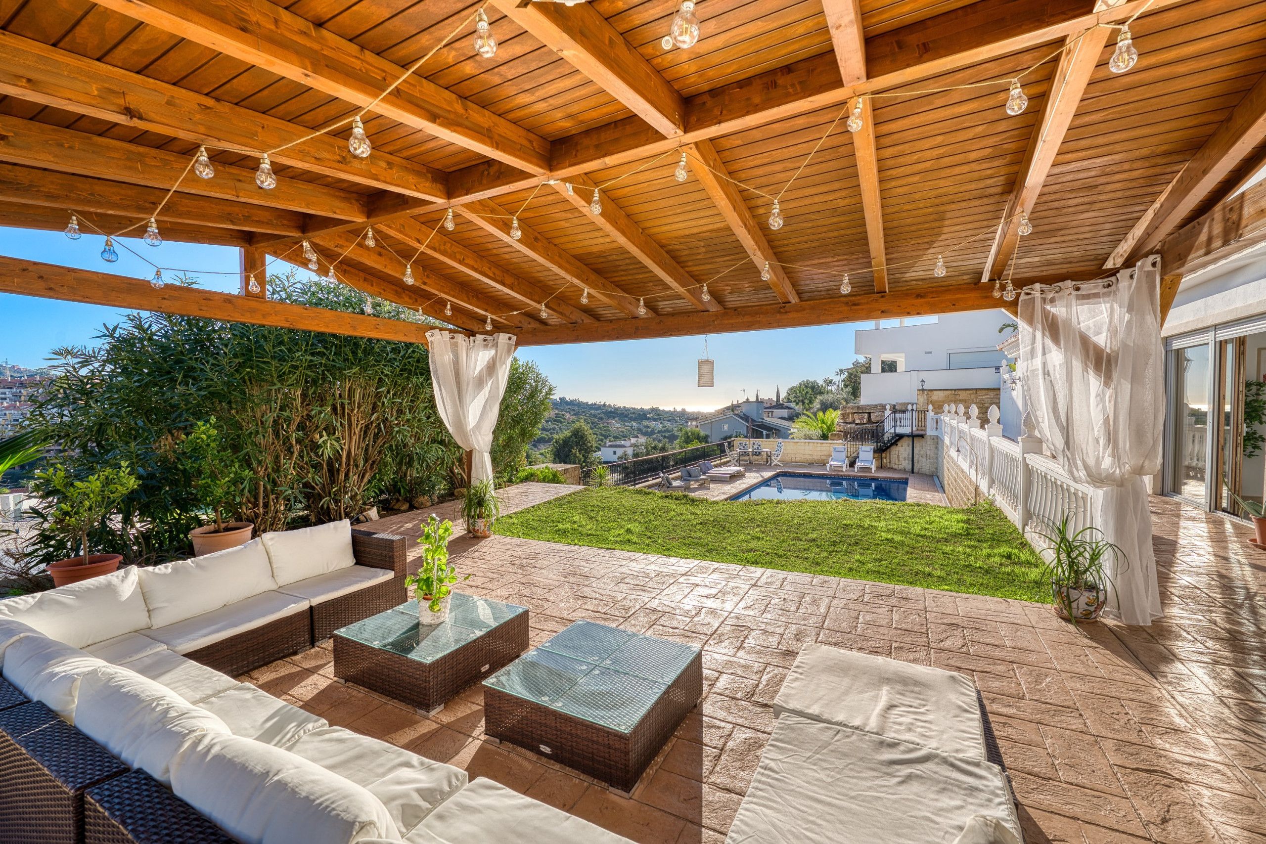 A covered patio with a wooden ceiling, outdoor furniture, and a view of a swimming pool and buildings in the background.