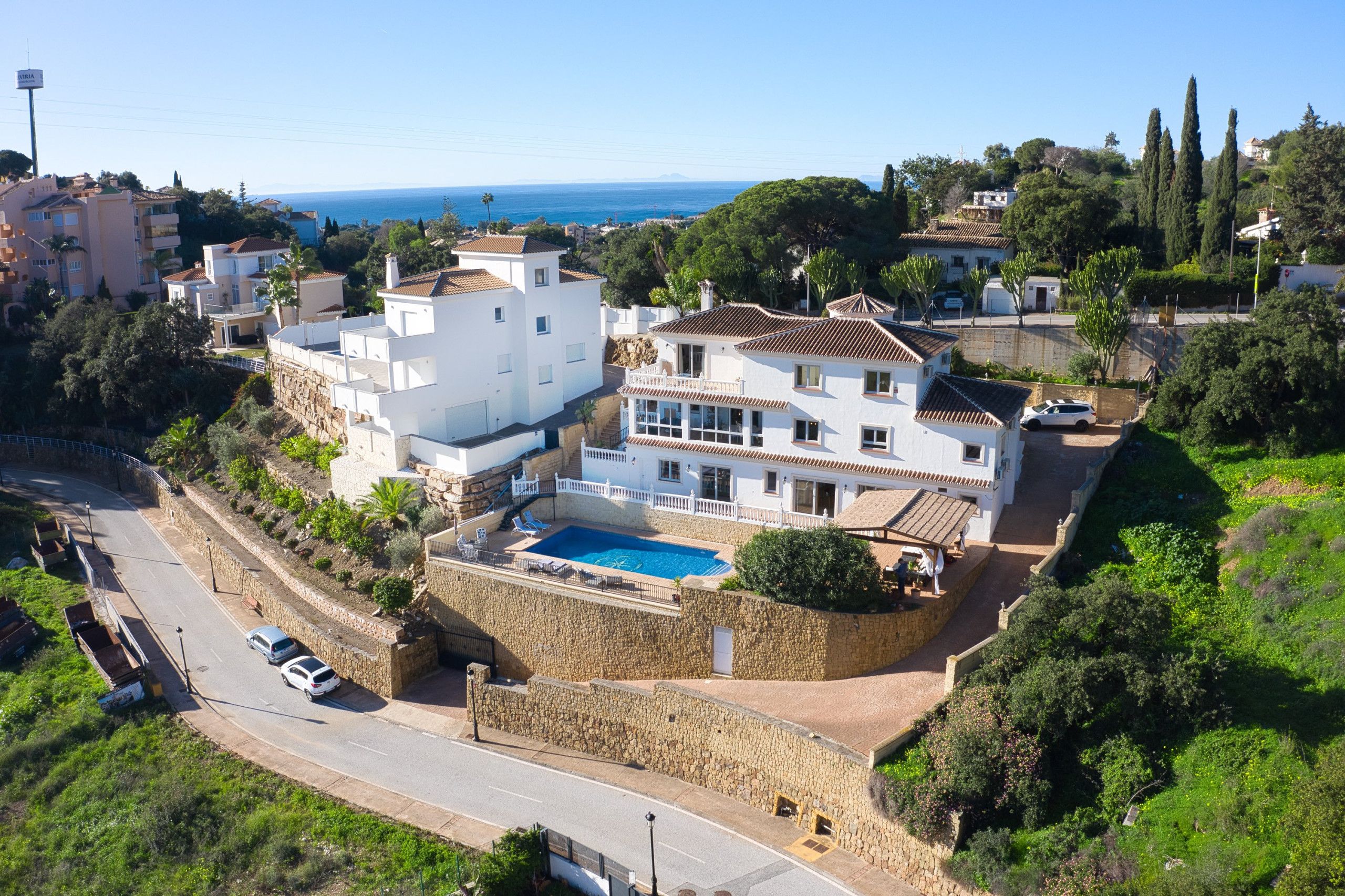 A coastal residential area with a large white house featuring a swimming pool, stone walls, and a view of the sea.