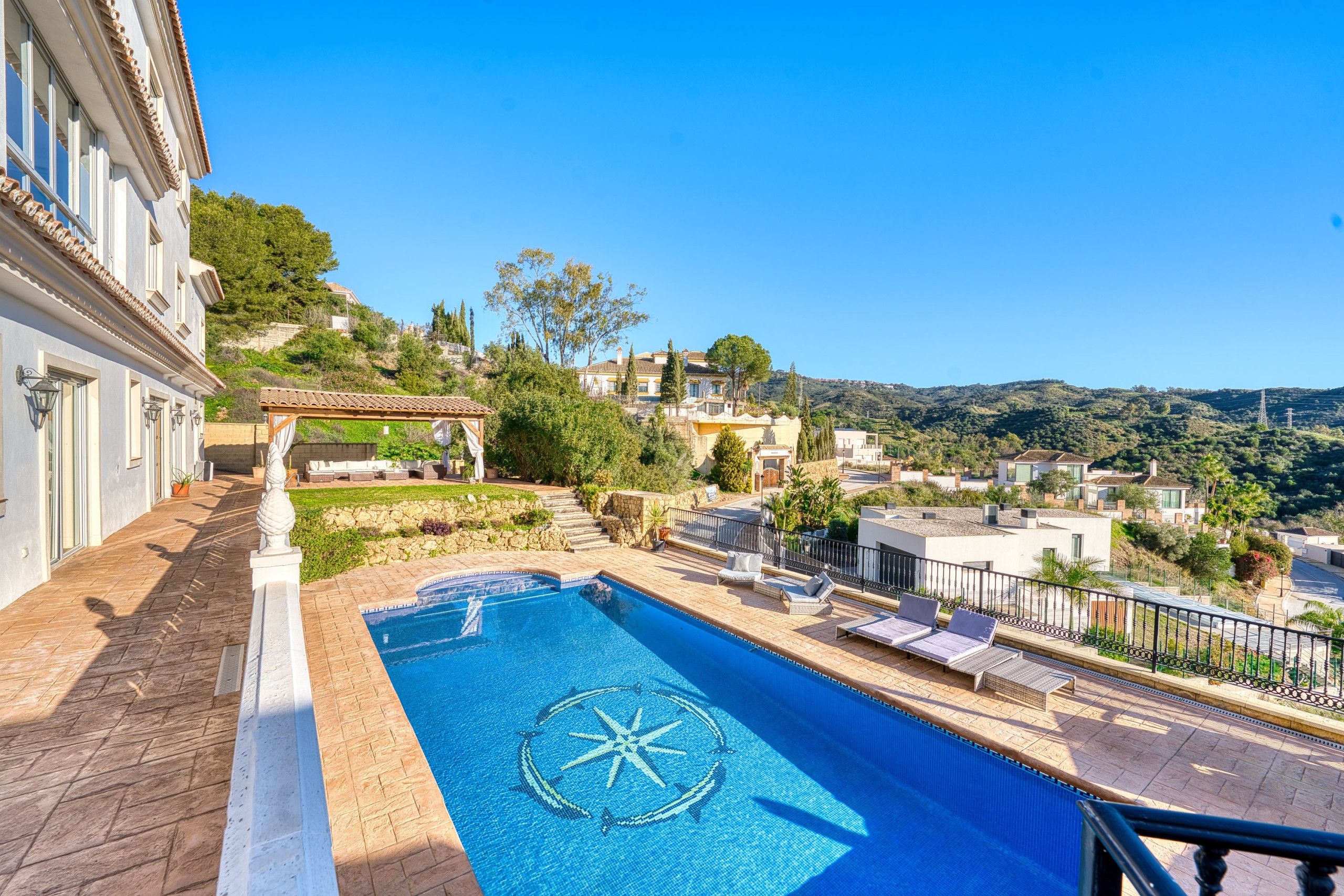 A swimming pool with a star design, surrounded by a terrace with lounge chairs and a view of a hillside residential area.