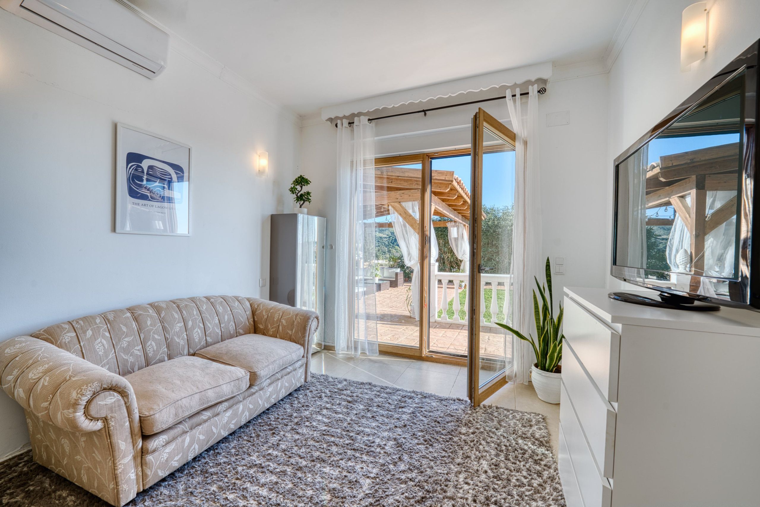 A modern living room with a beige sofa, a large flat-screen TV, and a sliding glass door leading to an outdoor patio.