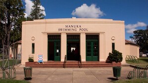 Un edificio con l'indicazione 'Manuka Swimming Pool', dotato di ingresso ben visibile, gradini di accesso e un cartello che segnala l'apertura.