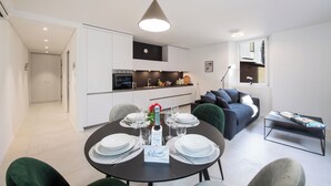 Ett modernt kök med vita skåp, en matplats med ett runt bord för fyra personer och ett vardagsrum med en grå soffa och ett soffbord.