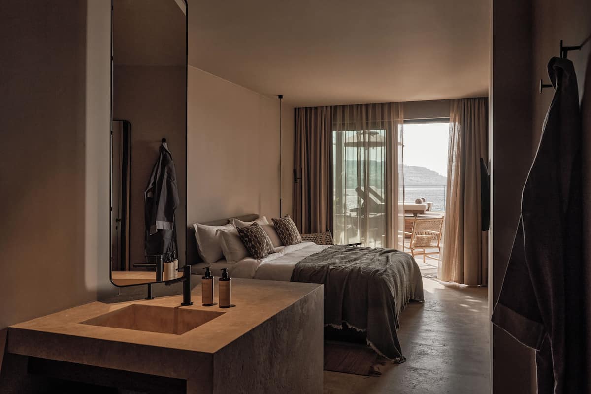 A modern hotel room with a large bed, a bathroom with a large mirror and sink, and a balcony with a view of the sea.