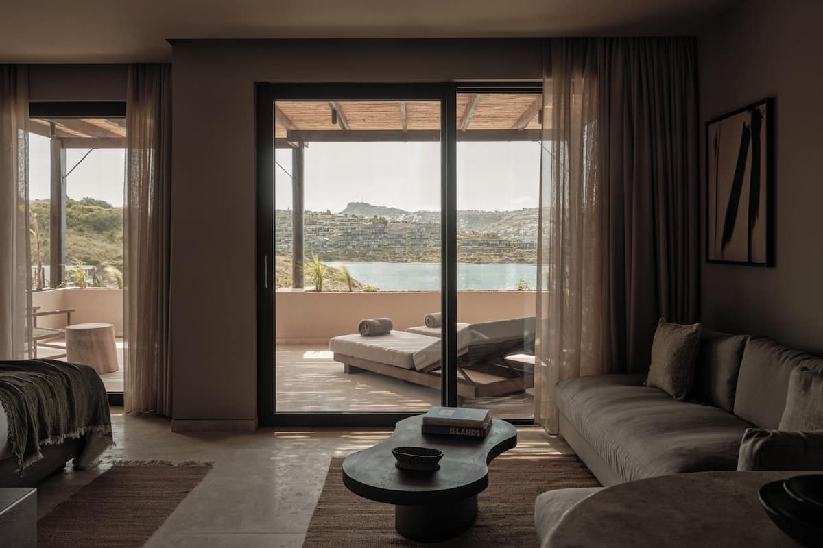 A modern living room with a sofa, a coffee table, and a view of a hillside and water through large glass doors.