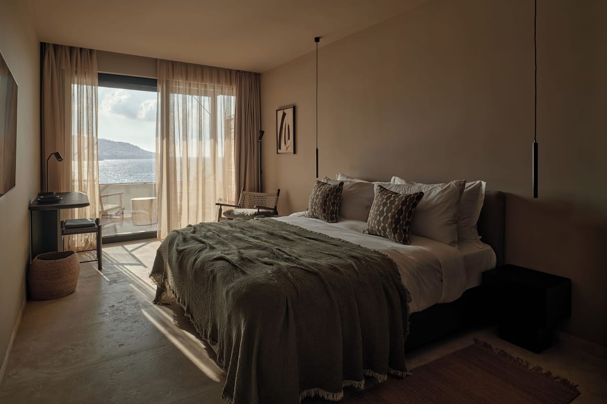 A bedroom with a bed, a chair, a desk, and a view of the sea.