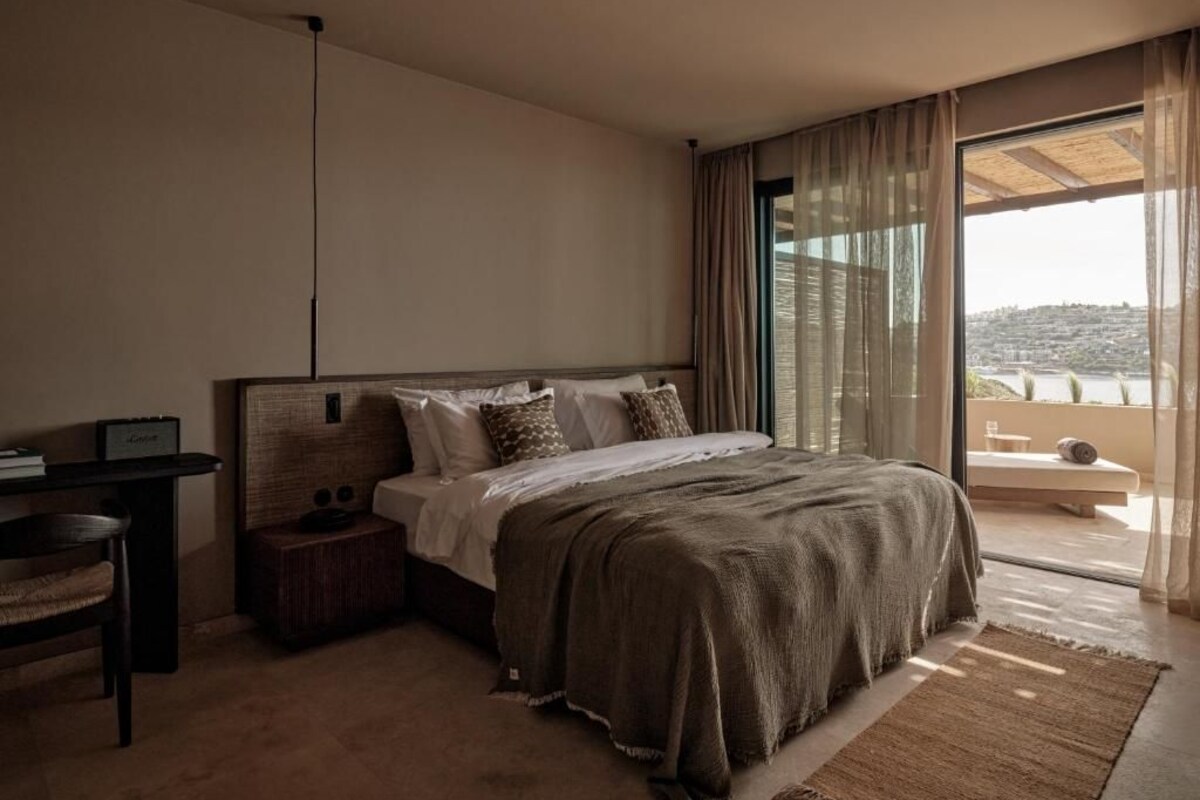 A bedroom with a large bed, a bedside table, a chair, and a view of the city through large windows.