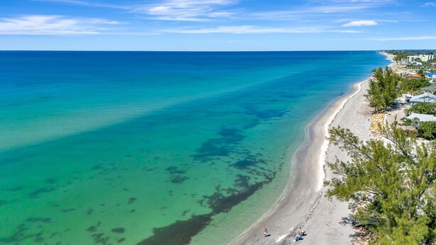 A coastal beach with clear turquoise waters, a sandy shore, and a few buildings along the coastline. - Steps to Beach | Pool + Hot Tub | 2025 Remodeled! (Englewood)