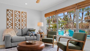 A living room with a grey sofa, two green armchairs, a wooden coffee table, and a large window with a view of palm trees and a pool. - Steps to Beach | Pool + Hot Tub | 2025 Remodeled! (Englewood)