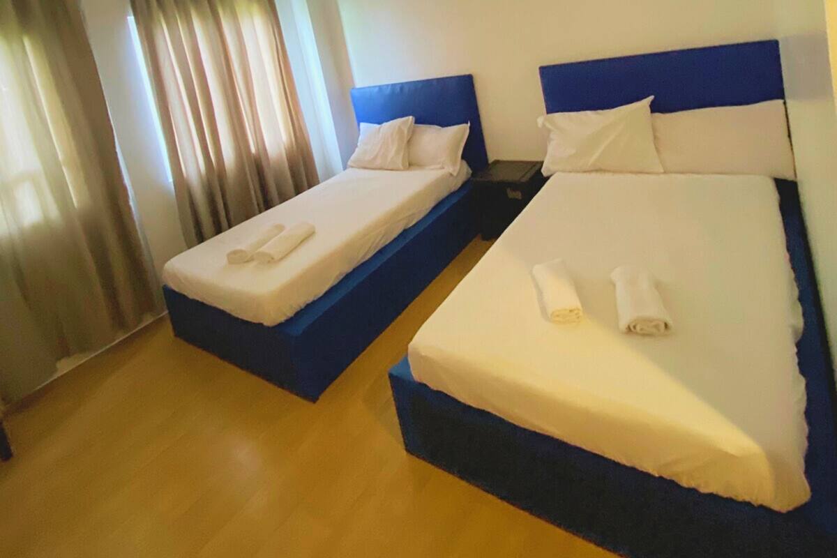 Two single beds with blue headboards and white linens, a wooden floor, and a window with curtains.