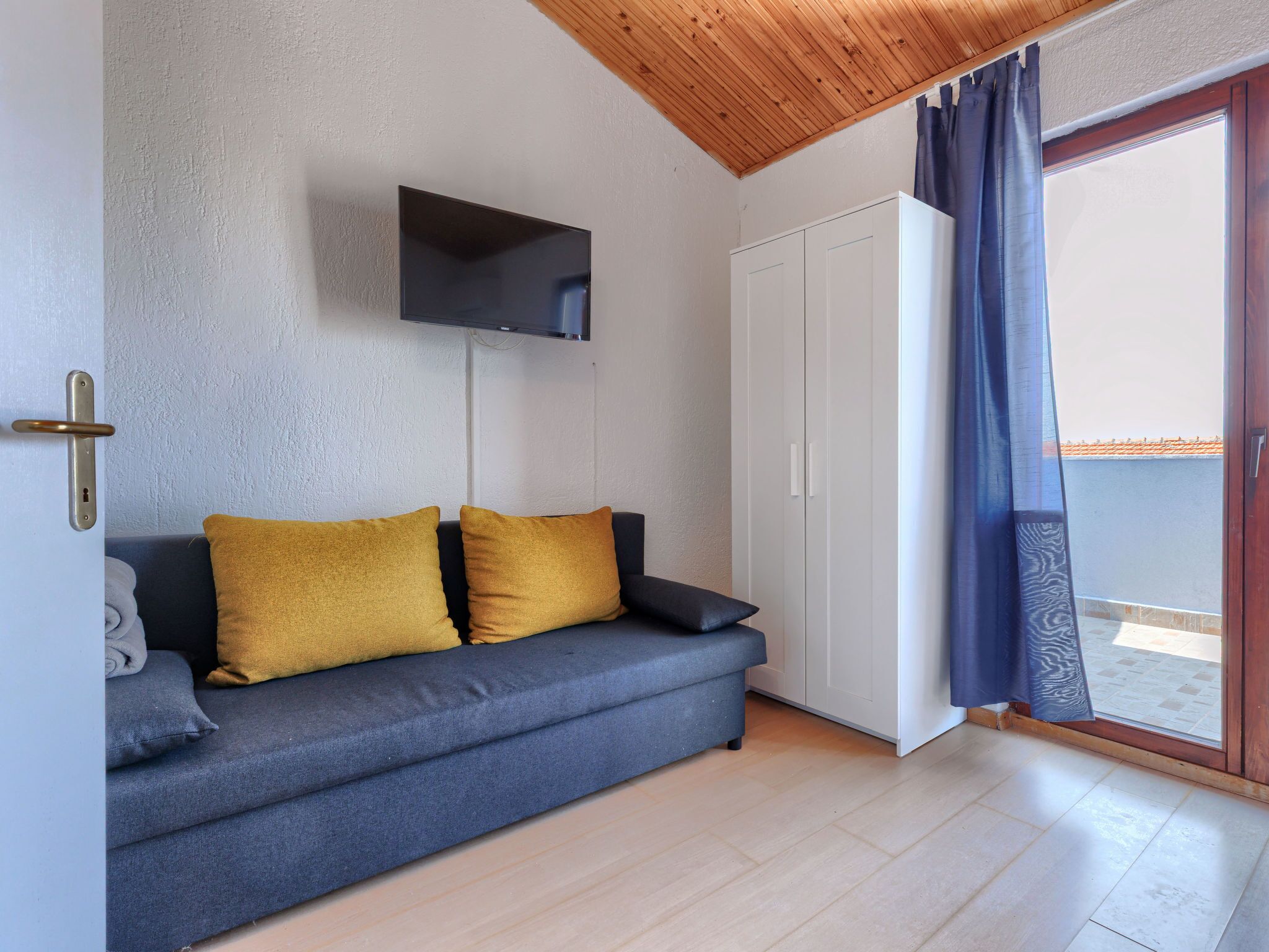 A room with a grey sofa, two mustard pillows, a flat-screen TV on the wall, a white wardrobe, and a sliding glass door with blue curtains.