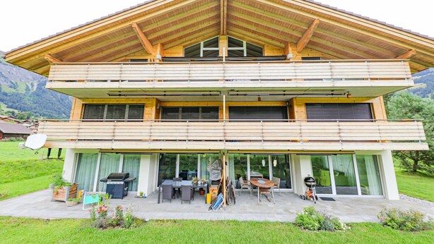 A two-story modern house with a wooden roof, large glass windows, and a patio area with outdoor furniture. - Kronenplatz 11 OG by Interhome (Lenk)