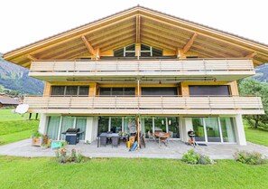 Een modern huis met twee verdiepingen, een houten dak, grote glasramen en een terras met buitenmeubilair.