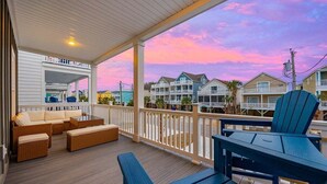 A covered outdoor deck with wicker furniture, a view of houses, and a sunset sky. - Luxury 5BR Escape | Pool & Hot Tub | Sleeps 16 (Surfside Beach)