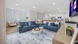 A modern living room with a blue sofa, a coffee table, and a kitchen in the background. - Luxury 5BR Escape | Pool & Hot Tub | Sleeps 16 (Surfside Beach)