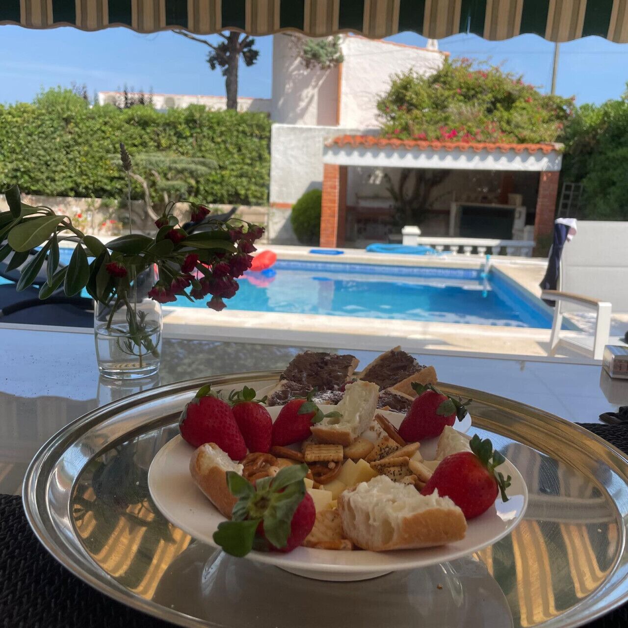 Une table à manger avec une assiette de nourriture et un verre d’eau, une piscine en arrière-plan et un bâtiment avec un toit rouge.
