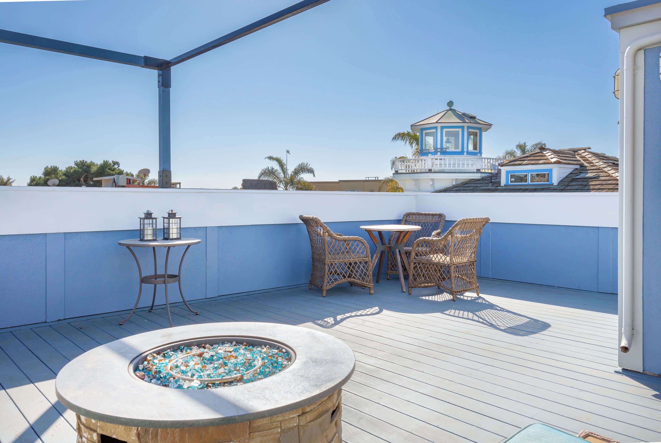A rooftop terrace with a stone fire pit, wicker furniture, and a view of a blue building with a tower.