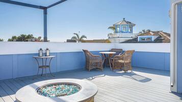 A rooftop terrace with a stone fire pit, wicker furniture, and a view of a blue building with a tower.