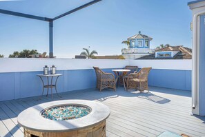 A rooftop terrace with a stone fire pit, wicker furniture, and a view of a blue building with a tower.