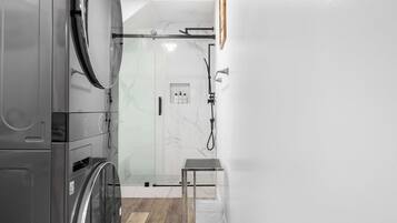 Un baño moderno con cabina de ducha de vidrio, lavadora integrada y una pequeña mesa.