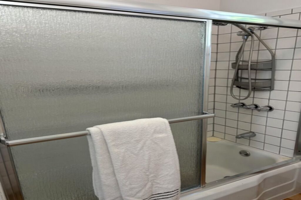 Un baño con ducha envuelta en vidrio, una toalla blanca colgada en un perchero y una bañera con ducha de mano.
