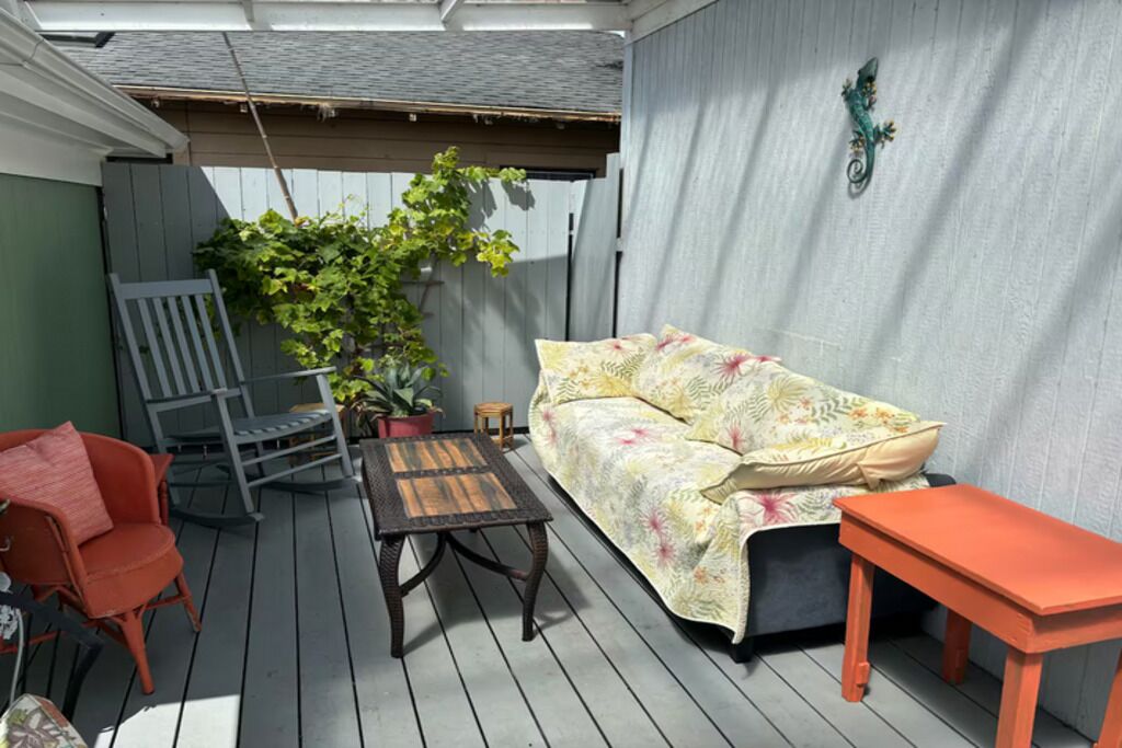 En overdækket terrasse med træterrasse, en grå stol, en sofa med blomstermønstrede puder, et lille bord og en potteplante.