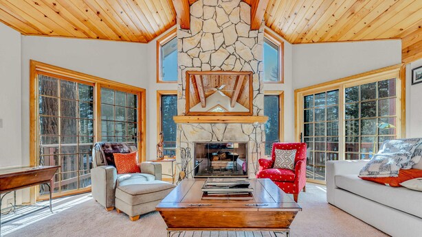 A spacious living room with a stone fireplace, large windows, and wooden ceiling beams.