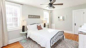 A bedroom with a bed, a ceiling fan, a bedside table with a lamp, a window with curtains, and a door. - Sojourn at Colley Manor Steps from Local Favorites Unit #6 (Norfolk)