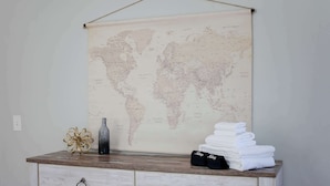 A world map on a wall, a wooden dresser with drawers, a decorative bottle, and folded towels. - Sojourn at Colley Manor Steps from Local Favorites Unit #6 (Norfolk)