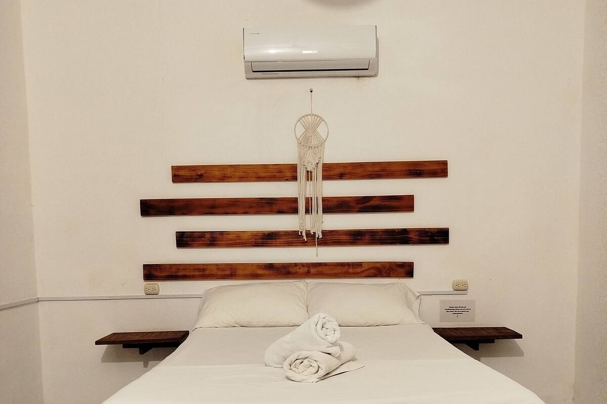 Une chambre à coucher avec une tête de lit en bois, un couvre-lit blanc, une table de chevet et un climatiseur fixé au mur.