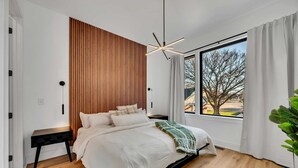 Un dormitorio moderno con un revestimiento de listones de madera, un ventanal con vistas, una cama con colcha blanca y una mesita de noche negra.
