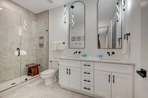 Een moderne badkamer met een glazen douchecabine, een wit toilet, een houten krukje, een grote spiegel en een witte wastafel met lades en een wastafel.
