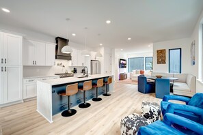 A modern kitchen with white cabinets, a central island with bar stools, and a dining area with blue chairs and a white sofa.