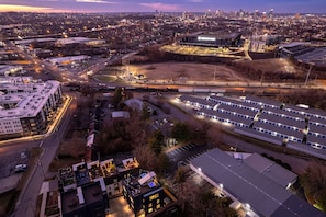 An aerial view of a cityscape at dusk with a large industrial complex, a parking lot, and a building with illuminated windows.