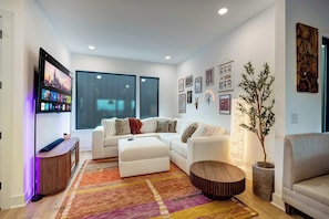 A modern living room with a sectional sofa, a round wooden coffee table, and a colorful rug. - Rooftop hot tub, sauna & game room house near Geodis Park, sleeps 12 (Nashville)
