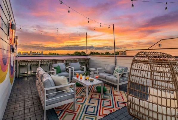 An outdoor seating area with a city skyline view at sunset, featuring a patterned rug, white furniture, and string lights. - Nashville-themed rooftop bar retreat, sleeps 12 near Geodis Park (Nashville)