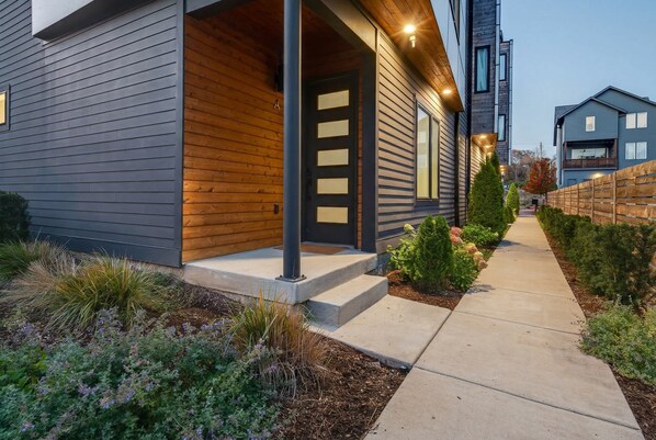 A modern two-story house with a wooden porch, a concrete pathway, and a well-maintained garden.