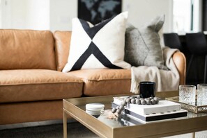 Un salon moderne avec un canapé en cuir marron, une table basse en verre et des objets décoratifs, notamment des livres, des bougies et un collier orné de perles.