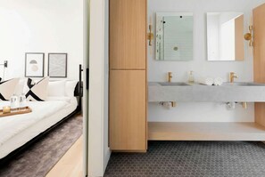 Habitación de hotel moderna con cama, armario de madera, baño con dos lavamanos y un espejo, y un suelo con estampado hexagonal.