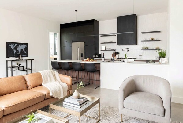 Una sala de estar moderna con sofá, sillón, mesa de centro y una zona de cocina con taburetes de bar.