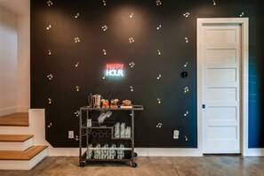 Una habitación con una pared negra que presenta un letrero de neón que dice 'HAPPY HOUR', un carrito de bar con vasos y botellas, y una puerta blanca.