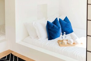 Cama bem arrumada com roupa de cama branca e almofadas azuis, um tabuleiro com velas e uma garrafa, e chão de madeira.