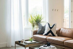 A living room with a leather sofa, a coffee table, a potted plant, and a window with sheer curtains. - Fall getaway: 2 rooftop homes with fire pits, sleeps 20, near The Gulch (Nashville)