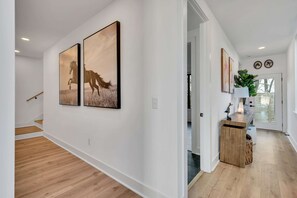A hallway with wooden flooring, white walls, and two framed pictures depicting horses. A wooden bench with a lamp and decorative items is visible. - Nashville house with hot tub, game room & pool table near Geodis Park (Nashville)