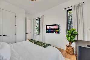 Quarto moderno com cama, televisão fixada na parede exibindo uma vista urbana, planta em vaso e armário.