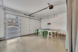 A clean, modern garage with a ping pong table and stools, a ceiling-mounted projector, and a garage door with multiple windows. - Nashville house with hot tub & pool table, sleeps 12 near Geodis Park (Nashville)