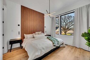 A modern bedroom with a wooden slat wall, a large window with a view, a bed with a white comforter, and a black nightstand. - Nashville house with hot tub & pool table, sleeps 12 near Geodis Park (Nashville)