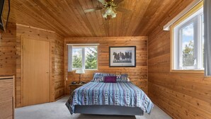 Un dormitorio con paredes de madera, una cama con ropa de cama estampada, un ventilador de techo y dos ventanas.