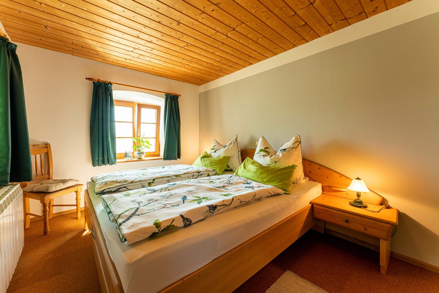 Un dormitorio con techo de madera, una cama con ropa de cama floral, una mesita de noche con lámpara y una ventana con cortinas.