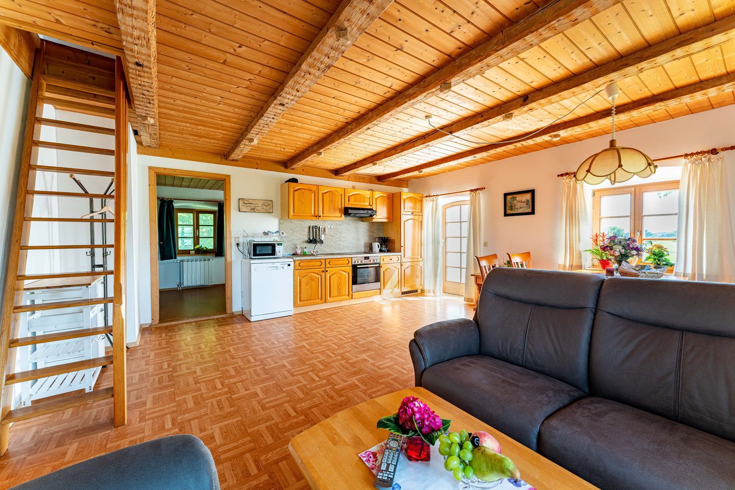 A spacious living area with a wooden ceiling, a sofa, a coffee table with fruits and a vase, and a kitchen in the background.